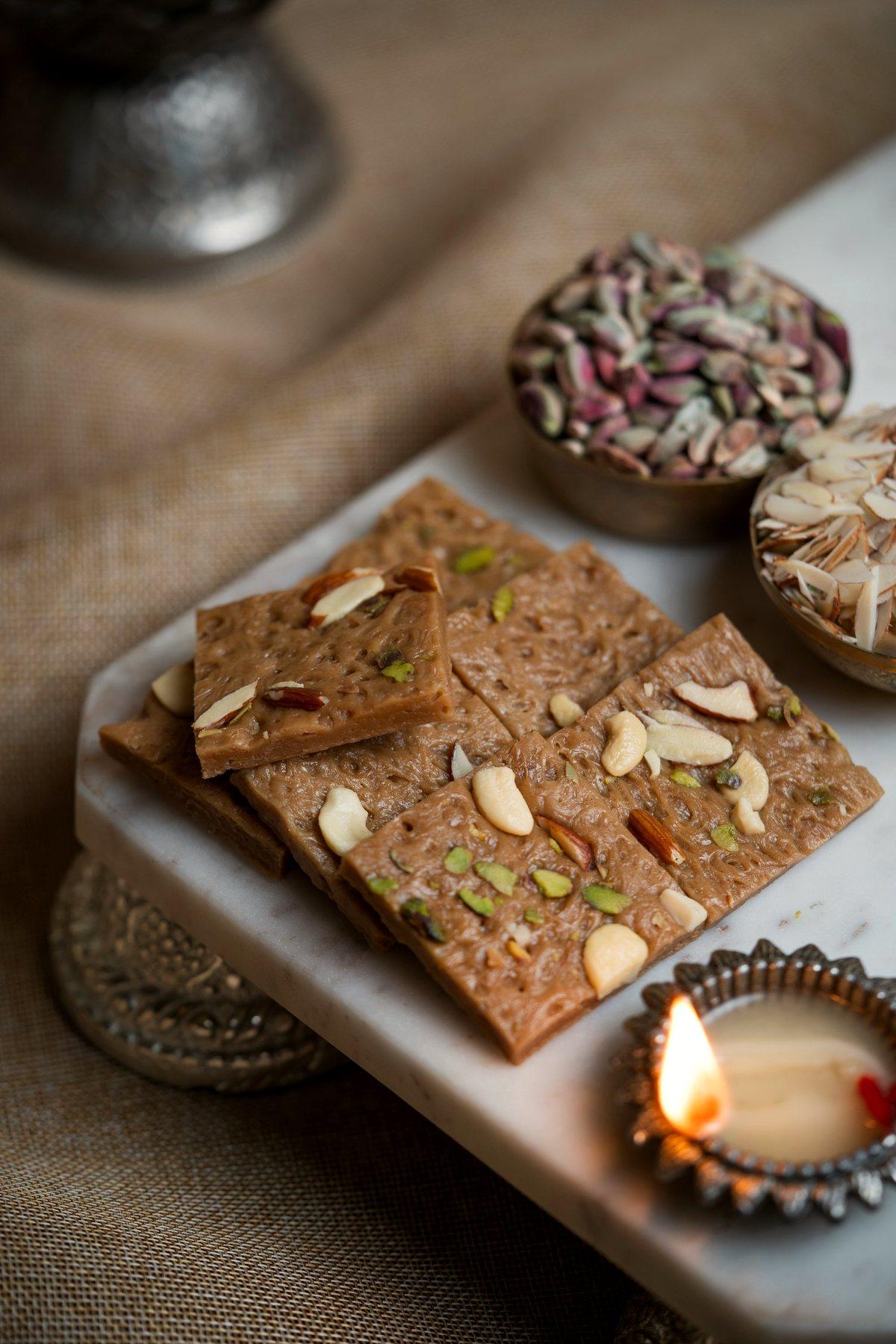 Slab of dry fruit barfi topped with sliced nuts beside a diya